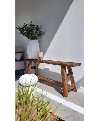 Een rustieke houten bank rust naast een witte muur, naast een grote vaas met het Kunst olijfblad 90 cm brandvertragend. Een rond wit kussen en een beige kaars voegen warmte toe en doen denken aan een mediterrane inrichting; gras verschijnt op de voorgrond.