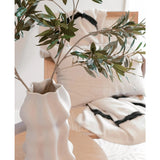 Een witte, sculpturale vaas met Kunst olijfblad 90 cm brandvertragend staat op een lichte houten tafel. Daarachter staan twee crèmekleurige kussens met zwarte strepen op een beige stoel, die knus comfort toevoegt aan deze moderne ruimte.