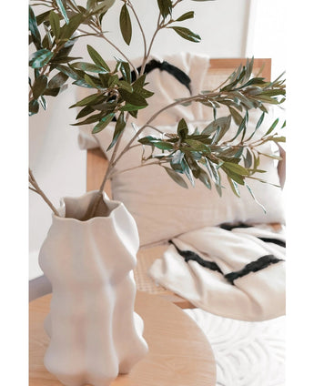 Een witte, sculpturale vaas met Kunst olijfblad 90 cm brandvertragend staat op een lichte houten tafel. Daarachter staan twee crèmekleurige kussens met zwarte strepen op een beige stoel, die knus comfort toevoegt aan deze moderne ruimte.