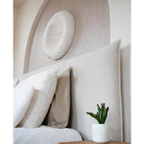 Een moderne slaapkamer heeft een beige gestoffeerd hoofdeinde, witte kussens, een Kunst vetplant 20 cm op een houten nachtkastje en ronde muurkunst met textuur boven het bed.