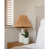 Een nachtkastje met een witte lamp met een geweven beige kap en de Kunst vetplant 20 cm staat naast een bed met wit en beige beddengoed, bij een raam met houten jaloezieën.