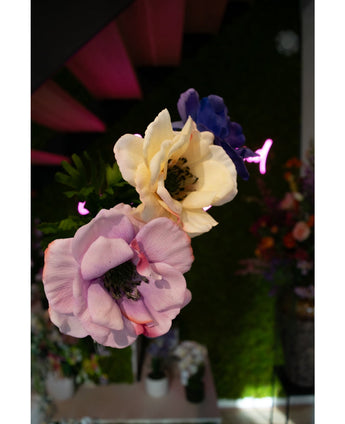 Close-up van Kunstanemoon Real Touch Blauw Paars 52cm kunstbloemen - lavendel, crème en blauw - tegen een vage achtergrond van groen, kleurrijke bloemen en roze neonverlichting.