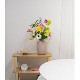 Een beige vaas met Kunstanemoon Real Touch paars 52cm bloemen staat op een licht houten plank naast een kleine ronde klok en een dienblad. Op de voorgrond staat een witte ronde tafel tegen een witte muur en gordijn.