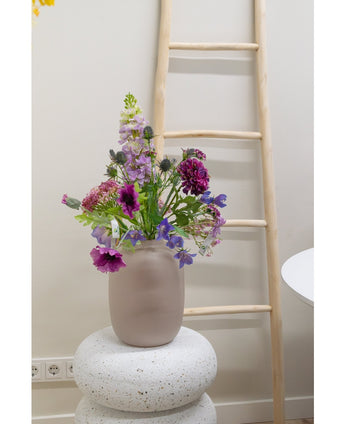 Een Kunstbloem Grasklokje van 62 cm paars staat in een matglazen vaas op een gestructureerde witte sokkel. Daarachter leunt een lichte houten ladder tegen een beige muur, met stopcontacten zichtbaar bij de vloer.