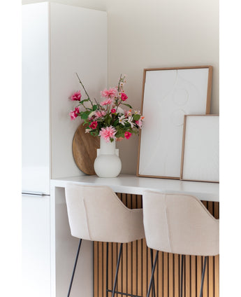 Een moderne witte keuken met twee beige barkrukken, een Kunstbloem Dahlia 68 cm roze in een witte vaas op het aanrecht en twee ingelijste abstracte kunstwerken tegen de muur.