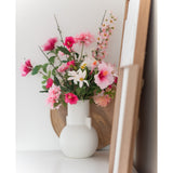 Een Kunstbloem Margriet 60 cm wit/roze staat in een witte vaas op een wit vlak naast kunst in houten lijst, met een rond houten dienblad zichtbaar op de achtergrond.