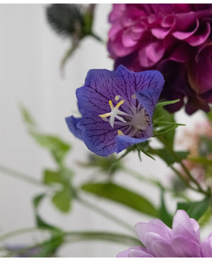 Een close-up van het Kunstbloem Grasklokje 62 cm paars, een delicate paarse kunstbloem met diepe nerven en gele meeldraden, tussen groene bladeren en zacht vage roze en paarse bloemen op de achtergrond.