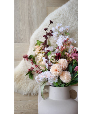De Kunstbloem Jilly 70 cm roze kunstbloem staat in een beige vaas op een houten vloer naast een zacht ivoorkleurig schapenvachtkleed.