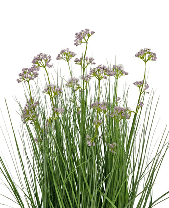 De Kunstgras Prei Bloem Ui Graspot 60cm heeft hoge, dunne groene bladeren en trossen kleine, delicate lichtpaarse bloemen op lange stelen. Het arrangement is brandvertragend en afgebeeld tegen een witte achtergrond.
