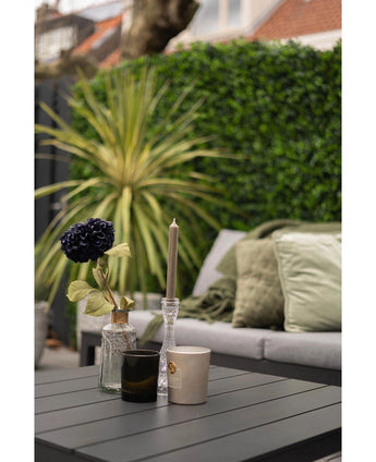 Een moderne buitentafel met een zwart oppervlak toont een vaas met een donkere bloem, een glazen kaarshouder en twee kopjes. Daarachter staat een bank met groene kussens tegenover een weelderige groene muur gemaakt van de Kunsthaag mattenmix 50x50 cm.