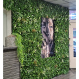 Een Kunsthaag Savanne brandvertragend paneel van 100x100 cm met weelderig groen omringt een groot zwart-wit gezichtsportret in een binnenruimte met betegelde plafondpanelen en een kleine toonbank aan de linkerkant.