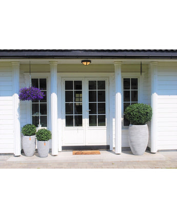 Voorportaal van een wit huis met dubbele deuren, zuilen en een hangende paarse bloemenmand. Drie Kunstplant Buxus op stam romeinse stijl UV 95cm plantenbakken met groene struiken staan aan weerszijden van de ingang.