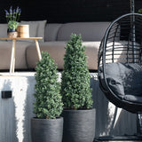 Twee Kunstplant Buxus Pyramide UV 120 cm staan op een houten dek voor een buitenzitje met een lichte bank, ronde tafel met lavendel en zwarte hangstoel.