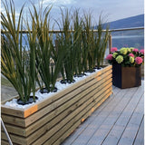 Een houten plantenbak met Kunstgras Groen UV 120cm en witte stenen staat op een balkon met glazen reling; vlakbij staat een zwarte vierkante pot met bloeiende roze, groene en witte hortensia's die uitkijken over het schilderachtige uitzicht.