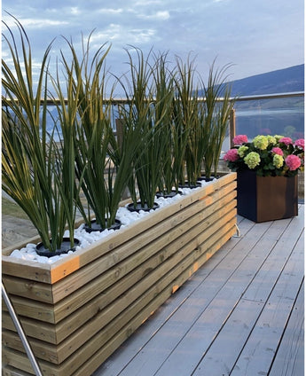 Een houten plantenbak met Kunstgras Groen UV 120cm en witte stenen staat op een balkon met glazen reling; vlakbij staat een zwarte vierkante pot met bloeiende roze, groene en witte hortensia's die uitkijken over het schilderachtige uitzicht.