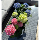 De Kunstplant Hortensia Groen UV 35 cm staat op een rieten tafel met glazen tafelblad naast een grijze buitenbank, met realistische groene hortensia's die oplichten in het zonlicht.