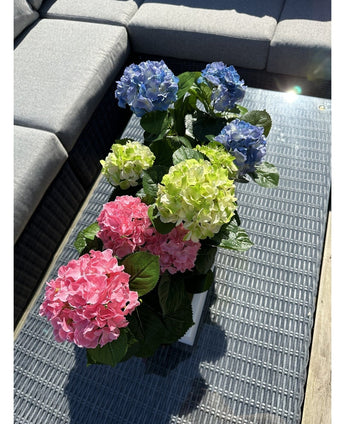 De Kunstplant Hortensia Groen UV 35 cm staat op een rieten tafel met glazen tafelblad naast een grijze buitenbank, met realistische groene hortensia's die oplichten in het zonlicht.