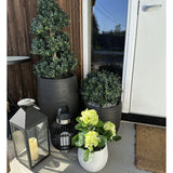 Een Kunstplant Hortensia Groen UV 35 cm in een witte pot met geelgroene bloemen staat op de veranda bij een bruine deur, naast twee planten in een zwarte pot en twee decoratieve lantaarns met kaarsen.