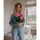 Een vrouw met krullend blond haar in een spijkerjasje en spijkerbroek staat binnen en glimlacht terwijl ze de Kunstplant Hortensia Roze UV 35 cm vasthoudt. Op de achtergrond staan een bank en een raam.