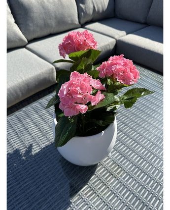 De Kunstplant Hortensia Roze UV 35 cm, een witte pot met levendige roze hortensiabloemen, staat op een geweven grijze buitentafel met een grijze sofa met kussens op de achtergrond, verlicht door zonlicht en zachte schaduwen.
