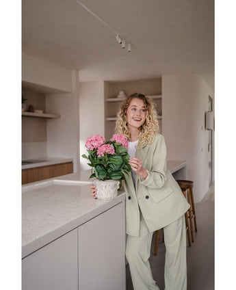 Een vrouw in een lichtgroen pak staat in een moderne keuken en glimlacht terwijl ze de Kunstplant Hortensia Roze UV 35 cm op het aanrecht vasthoudt.