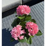 De Kunstplant Hortensia Roze UV 35 cm, met drie roze hortensiabloesems en groene bladeren, staat op een geweven grijze buitentafel, met een grijze bank met kussens op de achtergrond.
