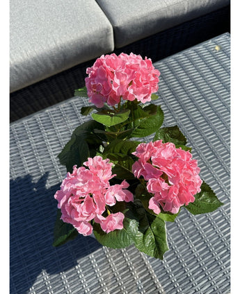 De Kunstplant Hortensia Roze UV 35 cm, met drie roze hortensiabloesems en groene bladeren, staat op een geweven grijze buitentafel, met een grijze bank met kussens op de achtergrond.