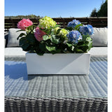 De Kunstplant Hortensia Roze UV 35 cm, een rechthoekige witte plantenbak met roze hortensia's, staat buiten op een rieten tafel met glazen blad, grijze kussens en een blauwe lucht op de achtergrond.