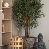 Een Kunst olijfboom UV 120 cm staat naast een grijs Boeddhabeeld en een houten lantaarn op de vloer, naast een lichte houten plank met handgevormde ornamenten. Deze onderhoudsarm plant brengt een rustige, serene sfeer in de ruimte.