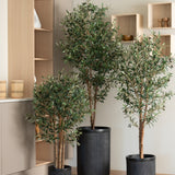 De kunst olijfboom UV 120 cm staat op een lichte houten vloer, met moderne licht houten planken en kasten op de achtergrond. De onderhoudsarme, UV-bestendige zwarte ribbelpot zorgt voor een eigentijdse look.