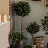 Een Kunst Olijfmontage UV 60 cm kunstmatige olijfplant met een ronde, bladerige top voegt onderhoudsvrij groen toe aan een moderne, minimalistische kamer, geplaatst in de buurt van houten planken en rieten manden.