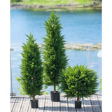 Drie Kunstplant Conifeer deluxe UV 120 cm bomen sieren een houten terras met glazen reling, met uitzicht op water en groen - ideaal voor elke kunstplant tuin dankzij hun UV-bescherming.