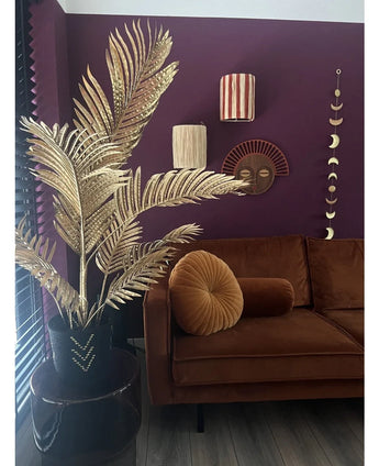 Een moderne woonkamer met een bruin fluwelen bank, rond plooikussen en de Kunstpalm Areca goud 120 cm in een zwarte vaas als gouddecor. Geometrische wandkunst hangt op een donkerpaarse muur, met houten vloeren en verticale lamellen in het zicht.