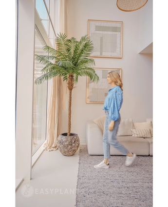 Een vrouw in een blauwe blouse en spijkerbroek loopt langs de Kunstpalm Phoenix De Luxe 210 cm bij een groot raam in een lichte, moderne woonkamer met beige muren, een bank en ingelijste kunst aan de muur.