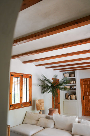 Een gezellige woonkamer met witte muren, houten balken, een witte sectionele bank, de Kunstpalm Phoenix De Luxe 210 cm, houten raam en deur, ingebouwde planken met manden en aardewerk, en natuurlijk licht dat door het raam naar binnen valt.