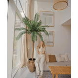 Een vrouw glimlacht met een drankje in een lichte, moderne woonkamer, staand naast de Kunstpalm Phoenix De Luxe 210 cm. De ruimte heeft grote ramen, een neutrale inrichting, ingelijste kunst, een bank, een houten tafel en geweven accenten.