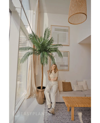 Een vrouw glimlacht met een drankje in een lichte, moderne woonkamer, staand naast de Kunstpalm Phoenix De Luxe 210 cm. De ruimte heeft grote ramen, een neutrale inrichting, ingelijste kunst, een bank, een houten tafel en geweven accenten.