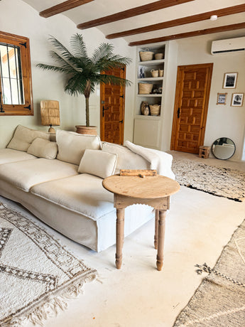 Een gezellige woonkamer met een witte bank, ronde houten bijzettafel, Kunstpalm Phoenix De Luxe 210 cm, gevlochten manden op planken, houten deuren, een ronde spiegel en neutraal getinte vloerkleden op een lichte vloer.