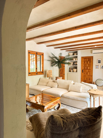 Een gezellige woonkamer heeft een grote crèmekleurige sofa, houten salontafel, Kunstpalm Phoenix De Luxe 210 cm in een pot, houten balkenplafond, ingebouwde planken met aardewerk, natuurlijk raamlicht en een warme, rustieke inrichting.