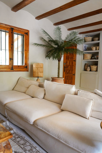 Een gezellige woonkamer met een grote crèmekleurige bank, de Kunstpalm Phoenix De Luxe 210 cm in de hoek, een houten lamp, ingebouwde planken met manden, houten plafondbalken en een raam met houten omlijsting.