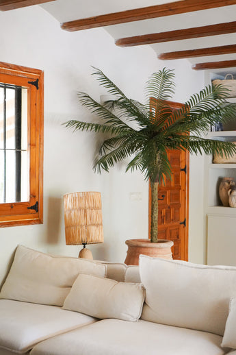 Een gezellige woonkamer met een witte bank, een rotan tafellamp, de Kunstpalm Phoenix De Luxe 210 cm in een pot, houten balken aan het plafond, een raam met houten kozijnen en een houten deur op de achtergrond.