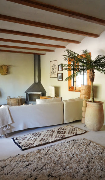 Een gezellige woonkamer heeft een witte bank, een vloerkleed met patroon, een houtkachel, ingelijste kunst en houten balken. Zonlicht stroomt naar binnen vanuit een raam naast de weelderige Kunstpalm Phoenix De Luxe 210 cm, waardoor de ruimte tot leven komt.
