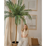 Een vrouw met blond haar die een beige topje en lichte broek draagt, glimlacht terwijl ze tegen de Kunstpalm Phoenix De Luxe 210 cm leunt in een lichte, moderne woonkamer met een neutrale inrichting en abstracte kunst aan de muren.