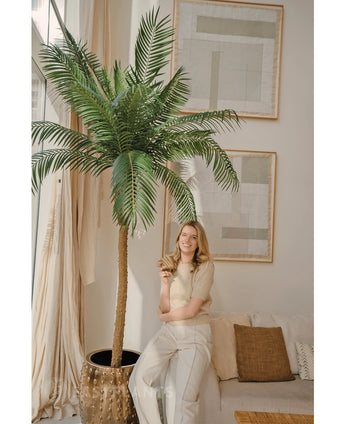 Een vrouw met blond haar die een beige topje en lichte broek draagt, glimlacht terwijl ze tegen de Kunstpalm Phoenix De Luxe 210 cm leunt in een lichte, moderne woonkamer met een neutrale inrichting en abstracte kunst aan de muren.