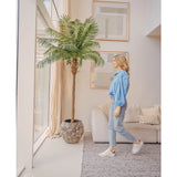 Een vrouw in een blauwe blouse en spijkerbroek loopt langs de Kunstpalm Phoenix De Luxe 250 cm in een lichte, moderne woonkamer met hoge ramen, neutrale inrichting en ingelijste kunst aan de muur.
