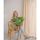 Een vrouw zit op een houten stoel en lacht terwijl ze de Kunstplant Alocasia UV 51 cm vasthoudt. Ze draagt een beige blazer, jeans en sneakers. Lichte gordijnen en een neutrale muur creëren een heldere, luchtige achtergrond.