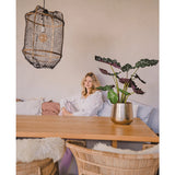Een vrouw in een witte blouse zit aan een houten eettafel met rieten stoelen. De gezellige, zacht verlichte kamer heeft de Kunstplant Alocasia UV 67 cm en een hanglamp met textuur als decor.