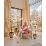 Een vrouw lacht op een beige fauteuil bij grote ramen met beige gordijnen, omringd door drie Kunstplant Bougainvillea 150 cm wit in een lichte, moderne leefruimte vol natuurlijk licht.