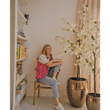 Een persoon zit glimlachend op een houten stoel naast de Kunstplant Bougainvillea 180 cm wit in een goudkleurige pot met textuur. Links staat een boekenplank met boeken en decor; daglicht stroomt naar binnen vanuit een raam aan de rechterkant.