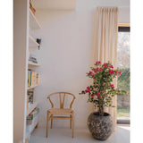 Een houten stoel staat naast een Kunstplant Bougainvillea Liana Fuchsia 115 cm, die een mediterrane flair geeft aan de lichte kamer. Boeken staan op een plank en beige gordijnen bedekken het zonovergoten raam.